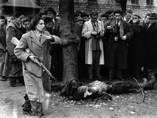 woman with rifle motioning others away from a dead body