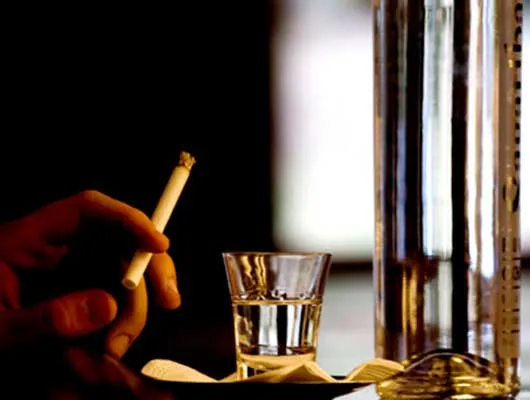 hand holding cigarette in front of drink in a glass and bottle